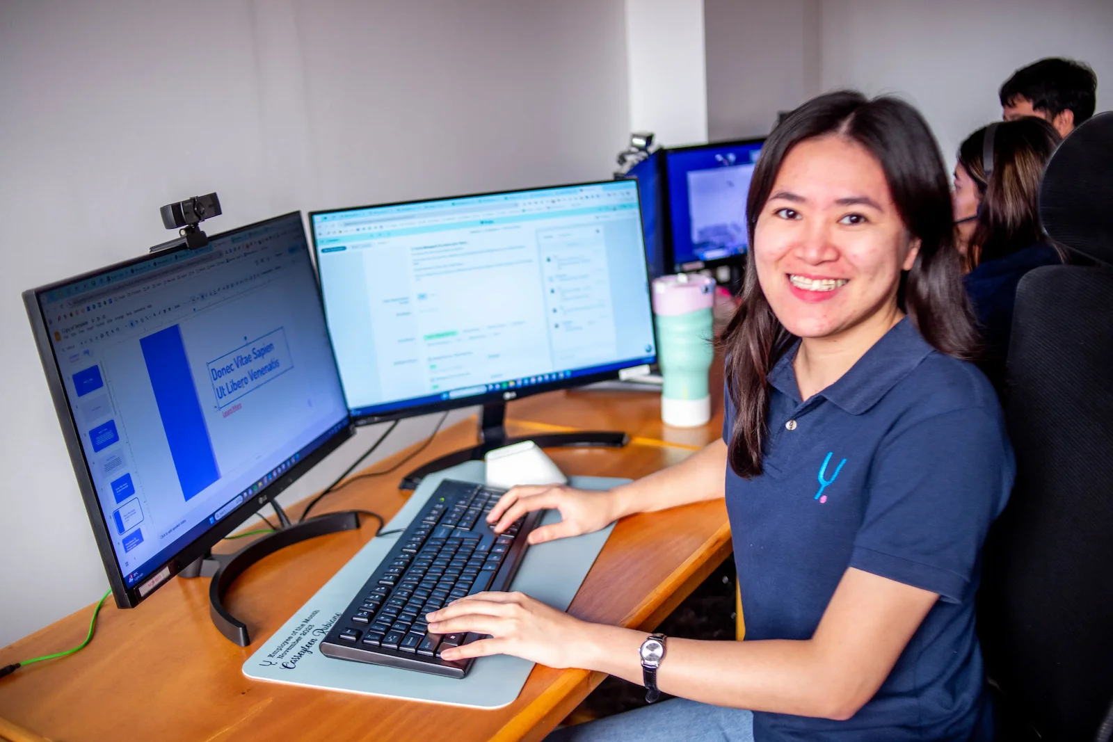 A woman smiles at her desk while working on a computer with two large monitors in a modern office.