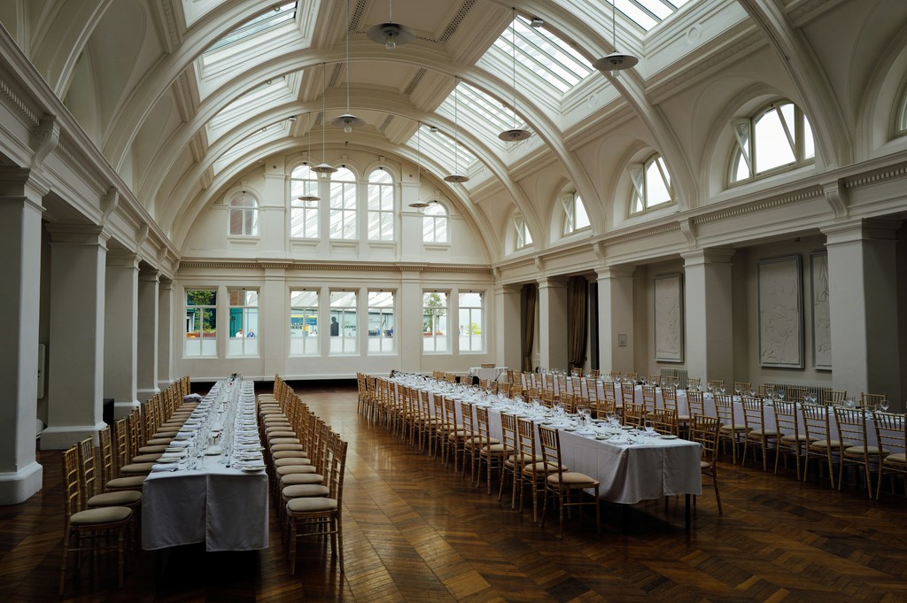 Grand wedding reception hall with arched skylights, white columns, long banquet tables, and gold chiavari chairs on a herringbone wood floor