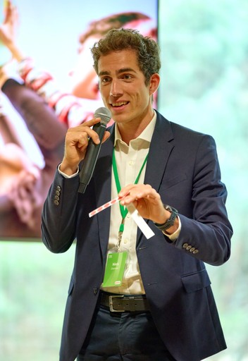 A young person with curly hair smiles at the camera while sitting in front of a laptop against a dark background.