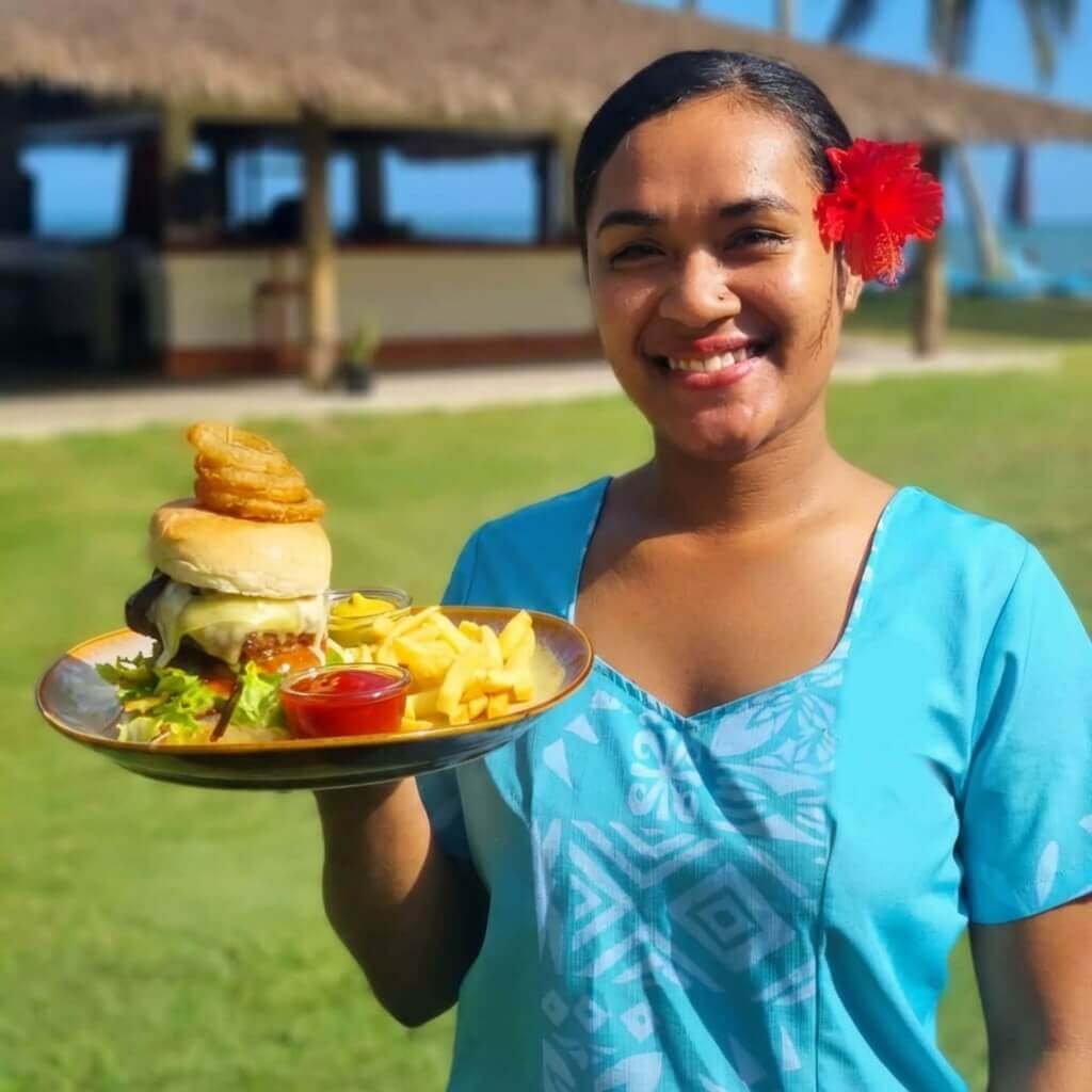 A female Fijian staff member holds a plate with a hamburger chips and salad at Uprising Resort Fiji