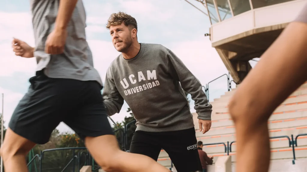 A focused individual wearing a UCAM University sweatshirt coaches athletes in a dynamic outdoor stadium setting, embodying the principles of the European Master in Sports Management.