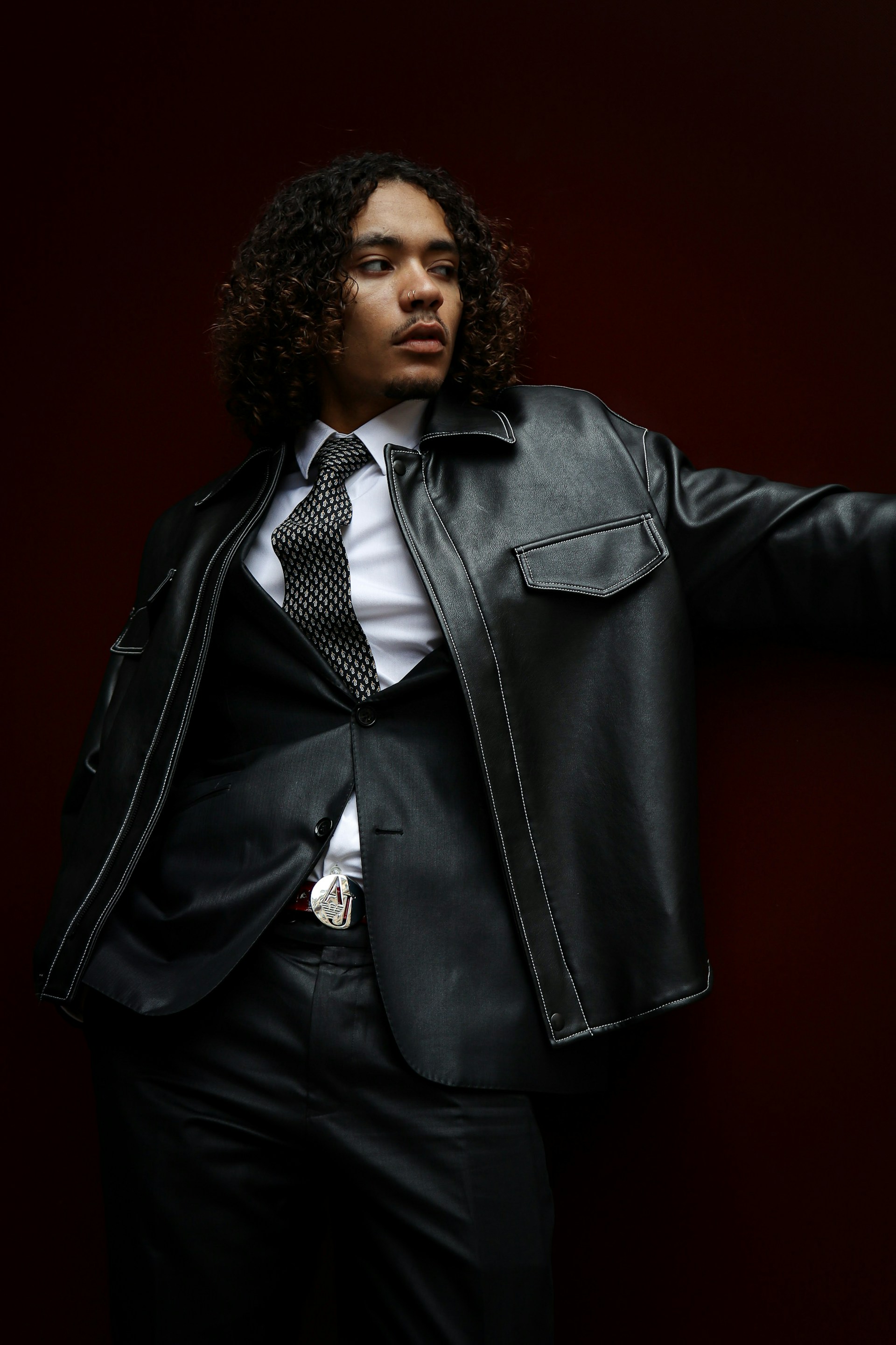 A low-angle shot of a man with curly hair looking off to the side while leaning against a dark maroon wall. He is dressed in a white shirt, a black tie, a black suit jacket, and a black leather jacket with visible white stitching.
