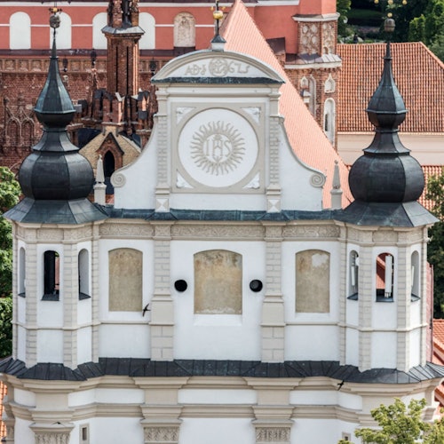 White Baroque-style church tower with two black domes, decorative details, and surrounding red-roofed buildings in the background.