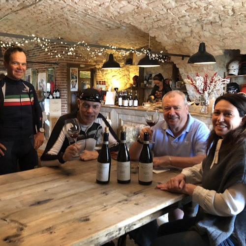 Cuatro personas están sentadas alrededor de una mesa de madera con botellas de vino, sosteniendo copas en una acogedora y rústica bodega de vinos adornada con luces de cadena.