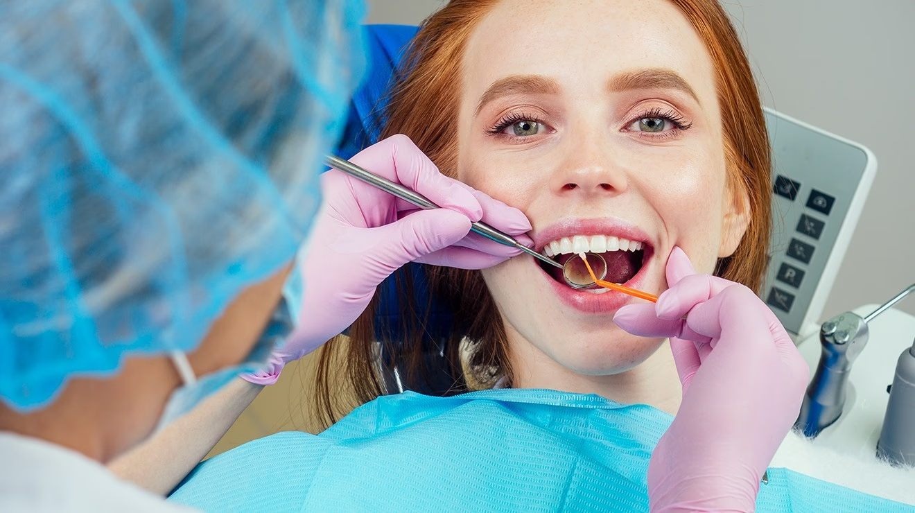 A young woman smiling brightly, showing perfect white teeth, while a dentist in gloves and a mask performs a Hollywood smile dental treatment at Bella Medical Centre. 