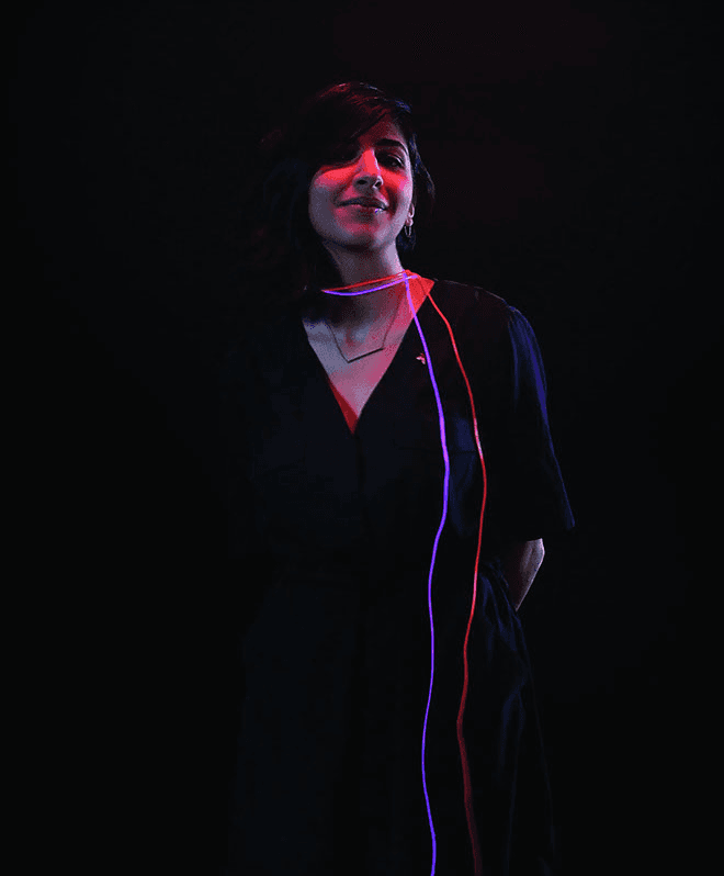 A person with shoulder-length dark hair stands against a black background, facing forward and smiling slightly. A thin, multicolored line runs vertically down the front of their dress, illuminated by red and blue light. The person’s right arm is slightly bent, and their left arm is behind their back. The lighting highlights the contrast between the dark background and the brightly lit line.