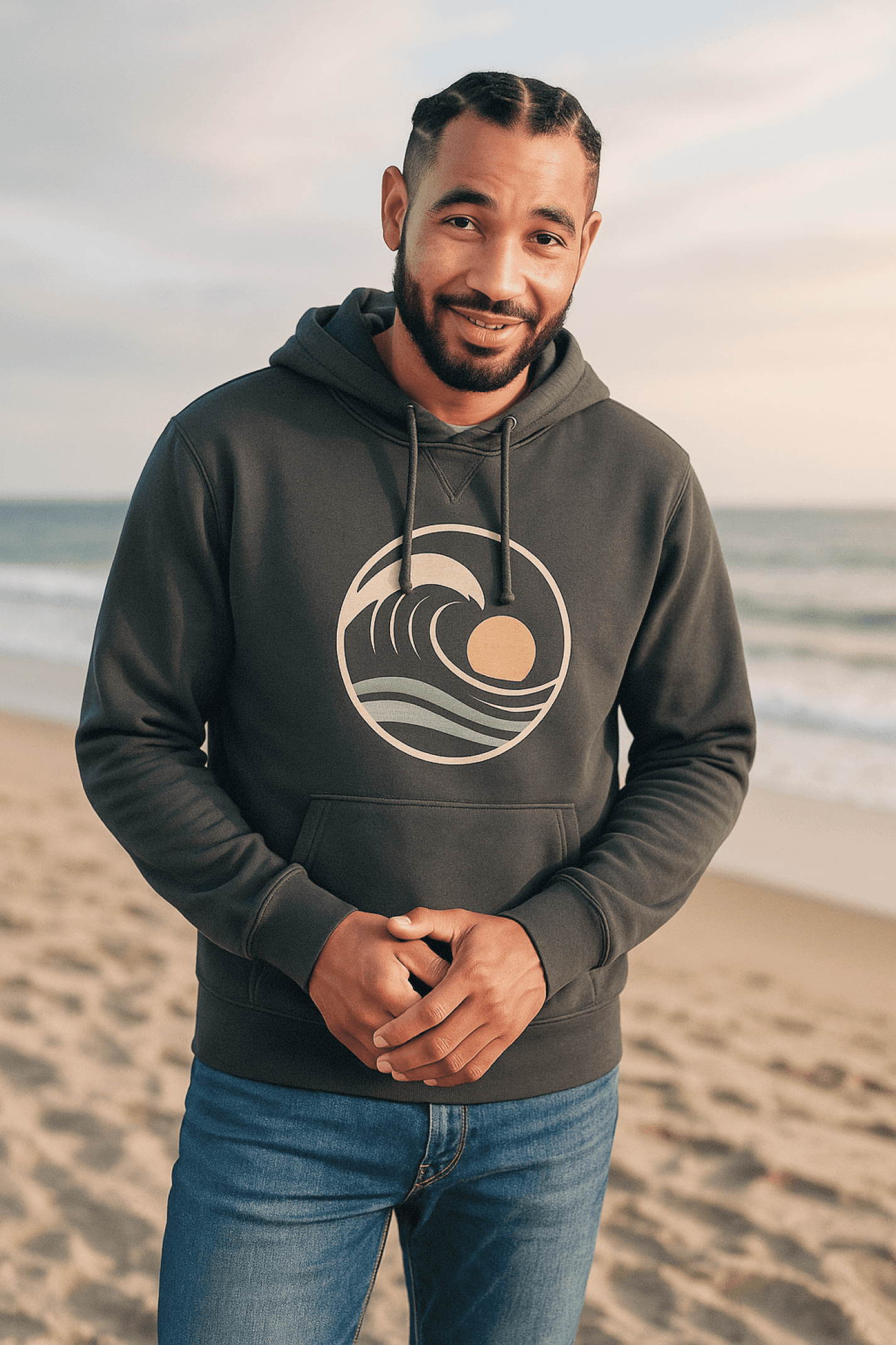 Man smiling, wearing a dark gray hoodie with a wave and sun graphic, standing on a beach.