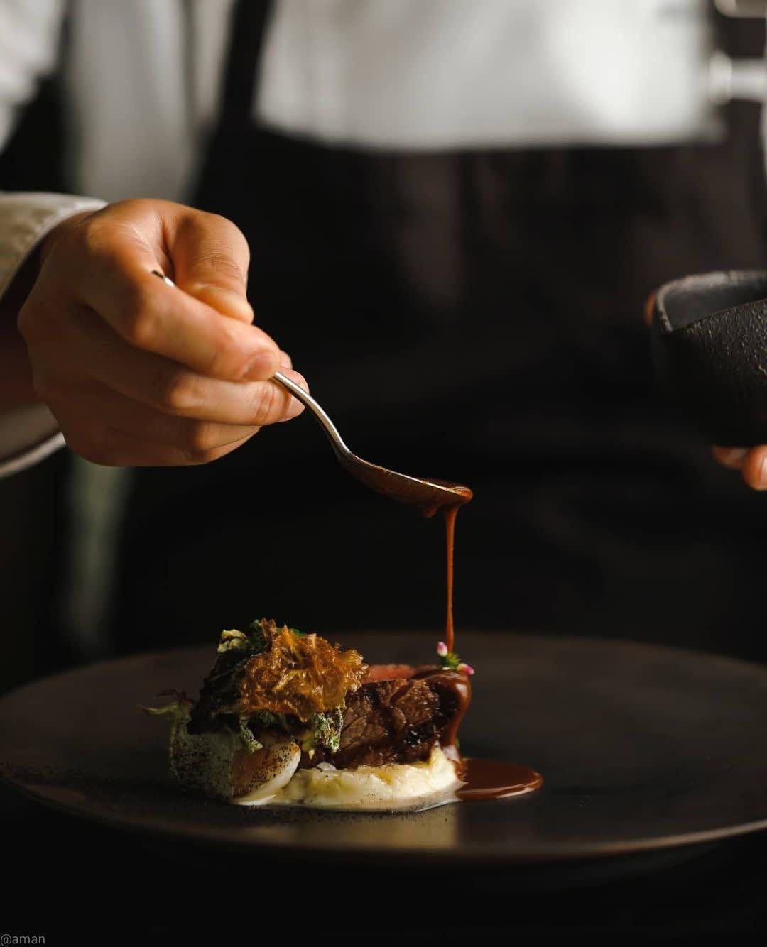 Chef finishing a refined dish with a sauce drizzle during evening service, capturing the warm, intimate bar-time atmosphere in Dhoom’s day-to-night dining transformation.