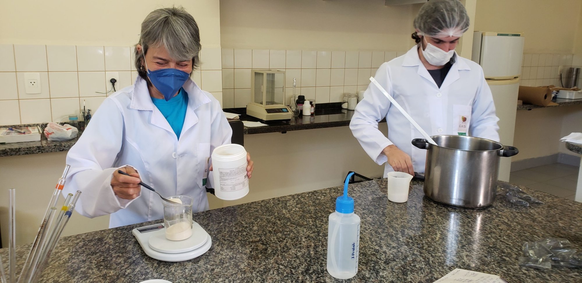 Duas pessoas de jaleco, touca e máscara manipulam frascos e panela em bancada de laboratório