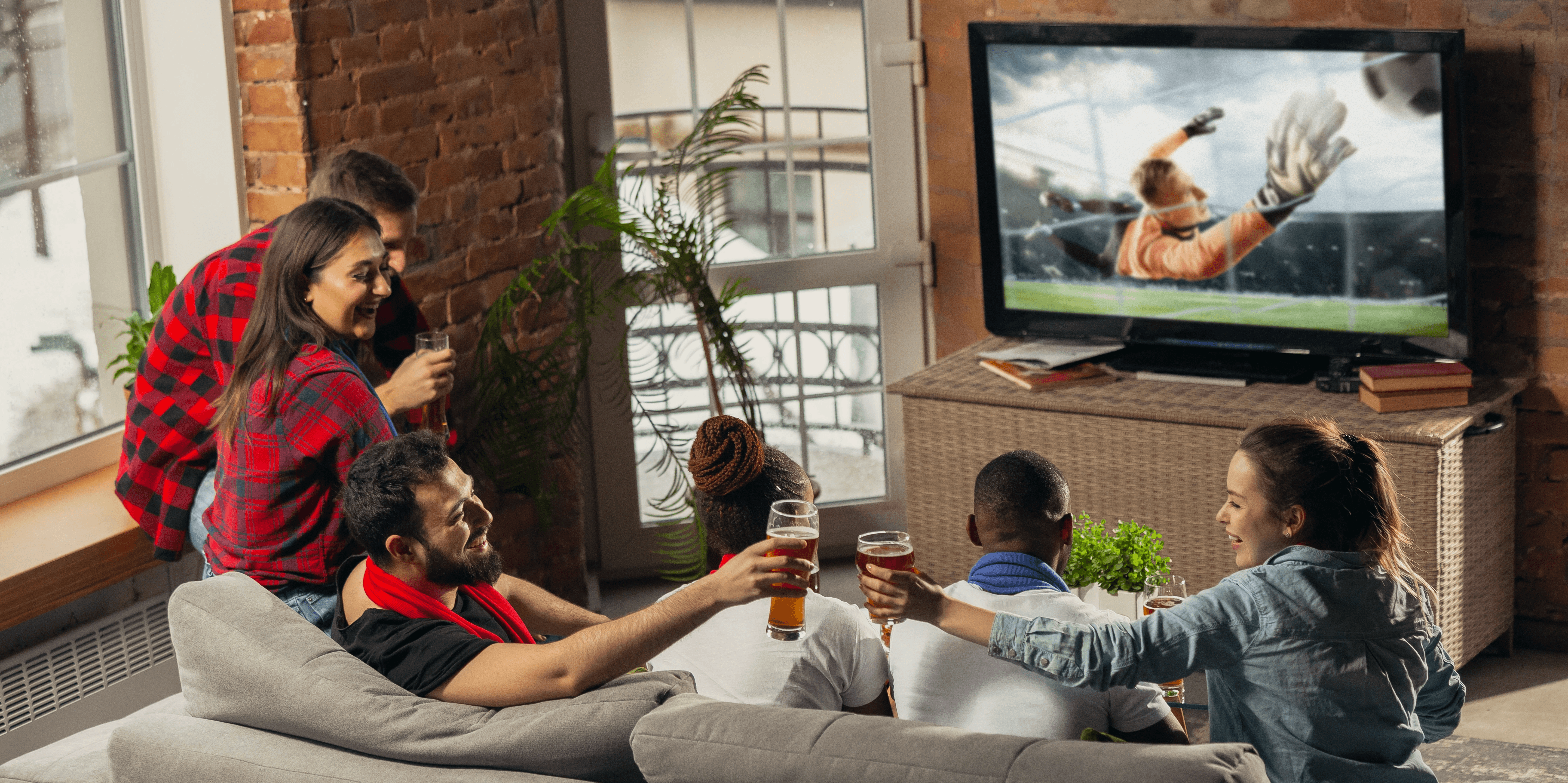 A group of friends gathers in a cozy living room with exposed brick walls to watch a soccer match on a large television, cheering and raising glasses of beer as a goalkeeper makes a dramatic save on screen. The diverse group is seated on a gray sectional sofa and nearby window ledge, creating a lively social atmosphere filled with plants and natural light. In the background, the scene transitions to the professional side of media and product development, featuring a digital platform for market research that uses advanced technologies like eye tracking and facial expression analysis to test consumer preferences for snack packaging and web layouts. Parallel to this digital analysis, a designer is shown at a bright workspace, meticulously sketching orange-themed mobile UI wireframes by hand, bridging the gap between social consumer experiences and the technical design process behind them.