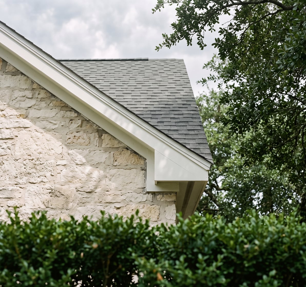 The gable end and new eave of a two-storey limestone house under a live oak canopy, with fresh fascia and new shingles