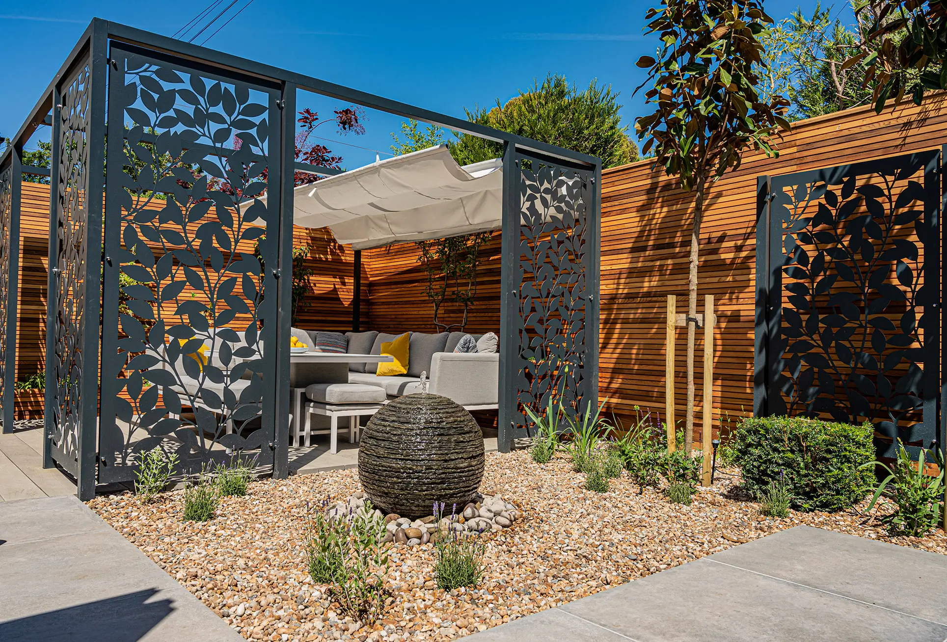 A modern outdoor space featuring decorative panels, landscaping, and a large stone sculpture. Clear blue sky above.