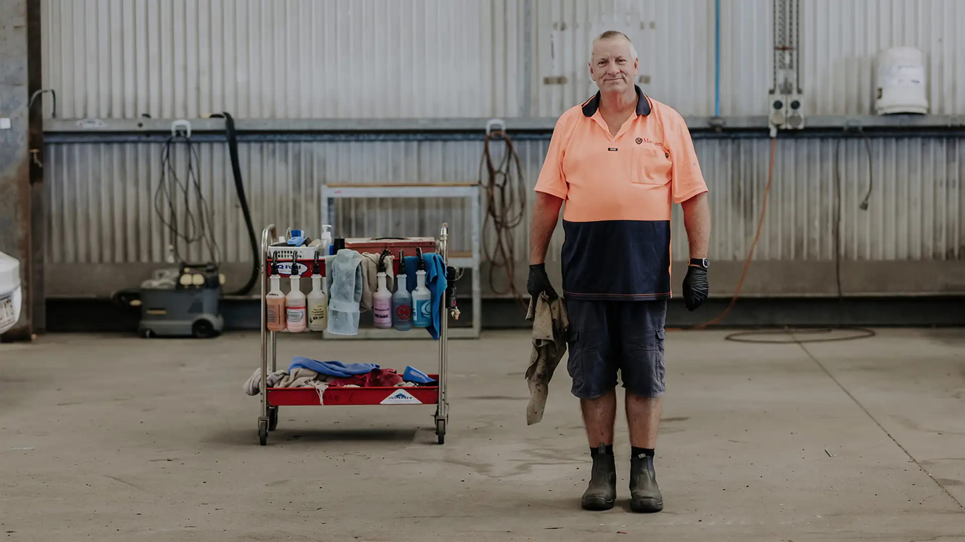 A Cox Automotive colleague standing inside a vehicle workshop beside a cleaning trolley, wearing high-visibility workwear.