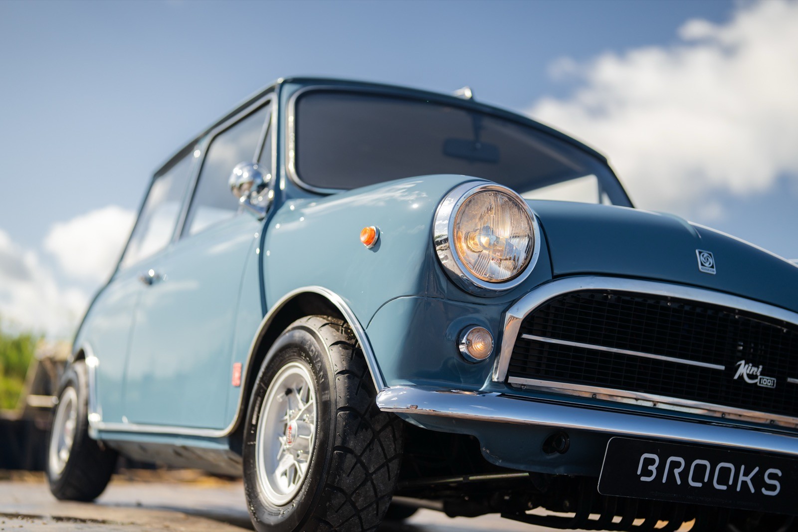 A classic blue Mini Cooper parked under a clear sky with fluffy clouds in the background.
