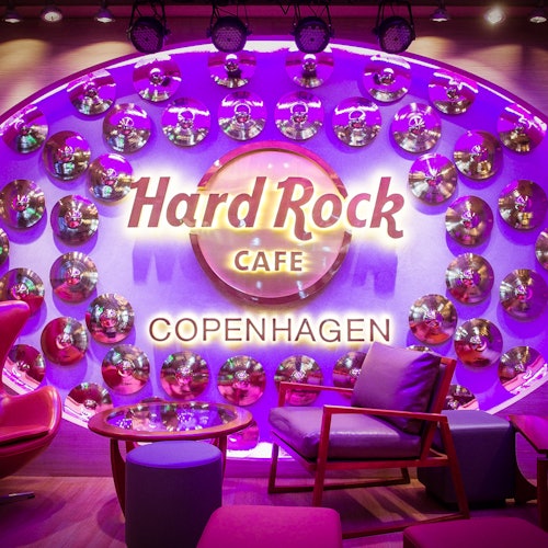Livligt siddeområde på Hard Rock Cafe København, med 'Hard Rock Cafe' skilt og dekorative spejldiske på en lilla-oplyst væg.