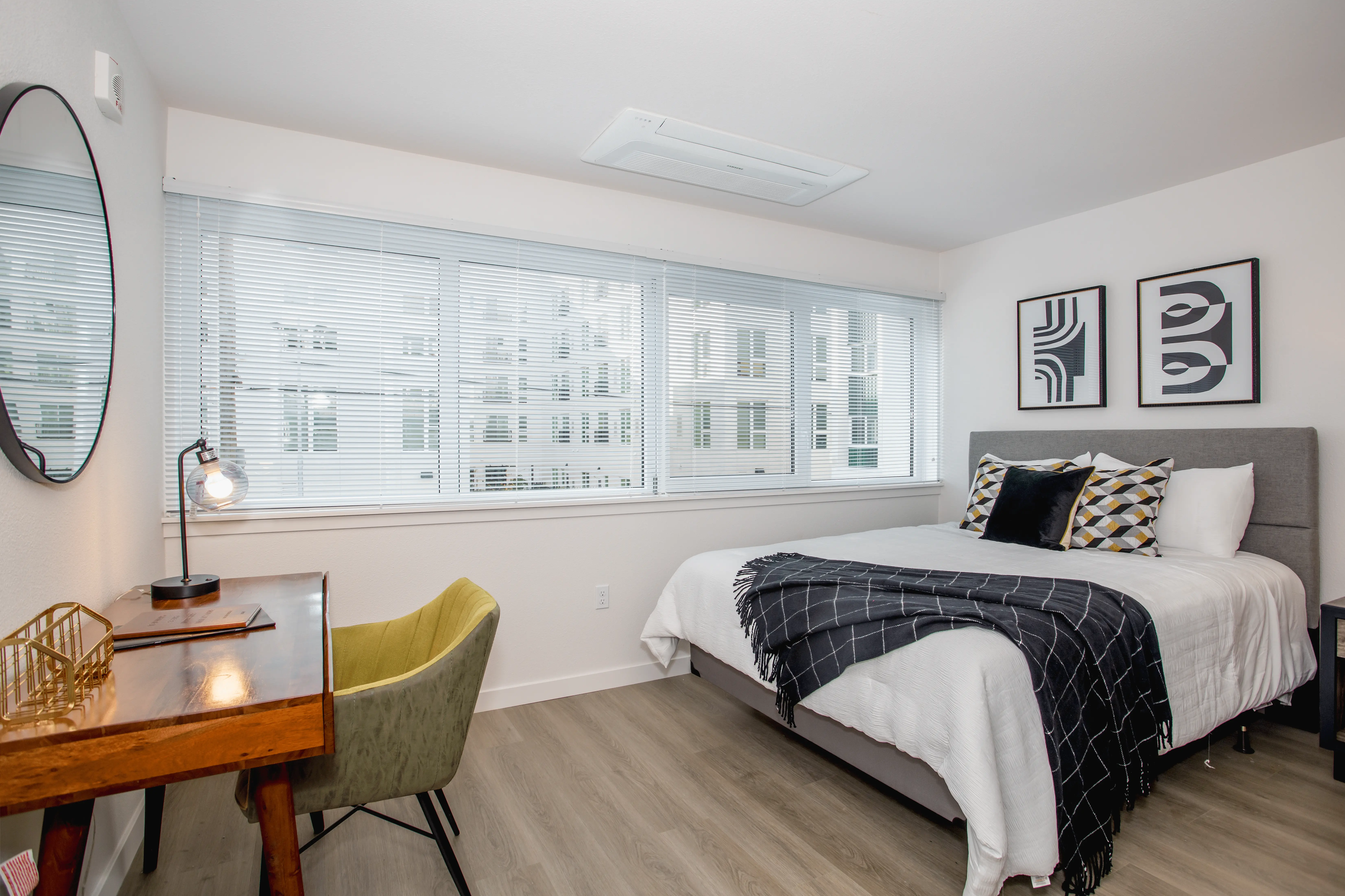 A modern bedroom in Midtown Sacramento features a large bed topped with a gray and black blanket and several decorative pillows. A wooden desk with a lamp and a green chair is placed beside it. The sun-drenched room boasts wide windows with blinds and framed abstract art on the wall.