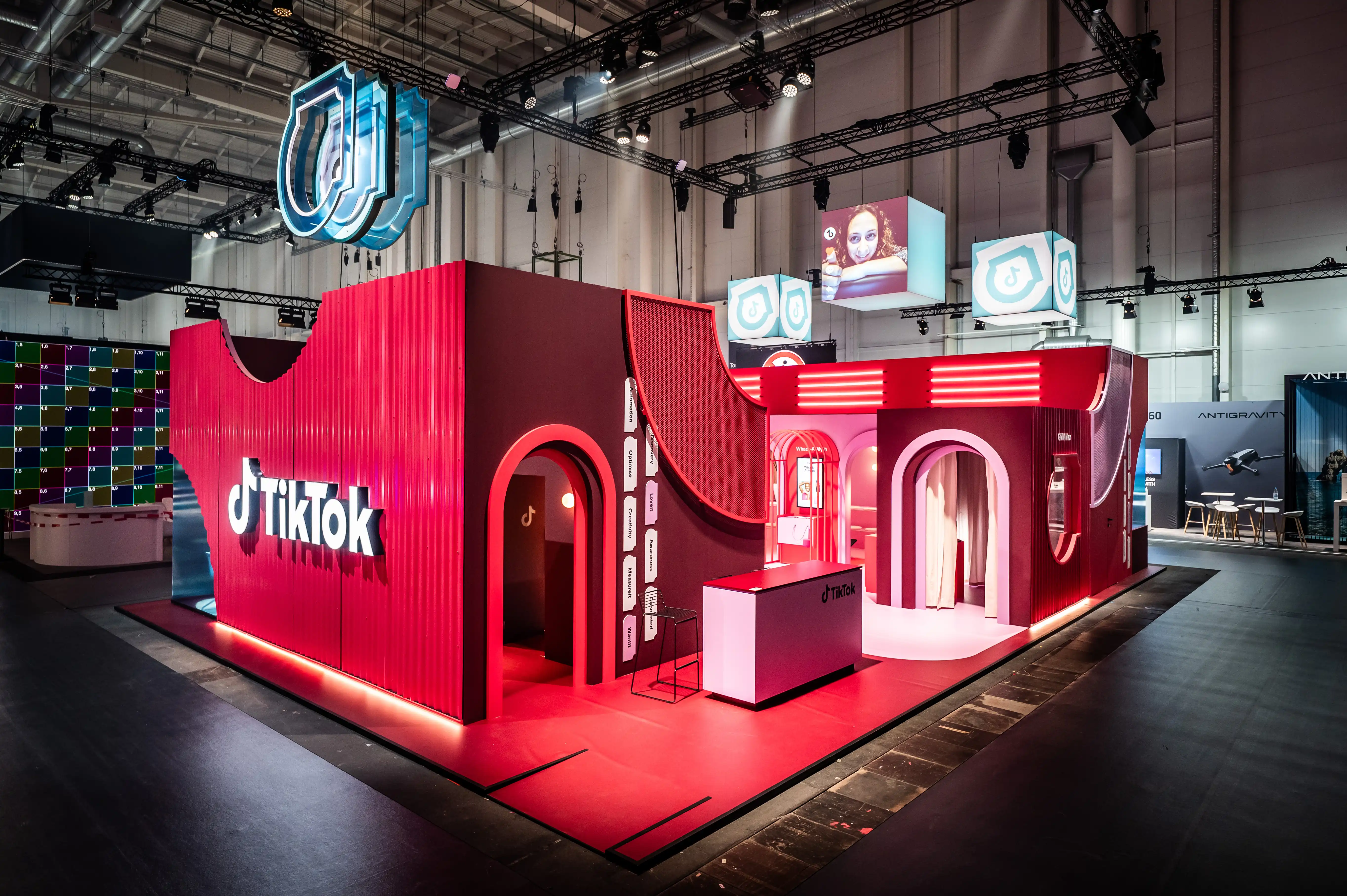 Red TikTok booth with curved facade, arched entrances and floating logo cubes in the exhibition hall.