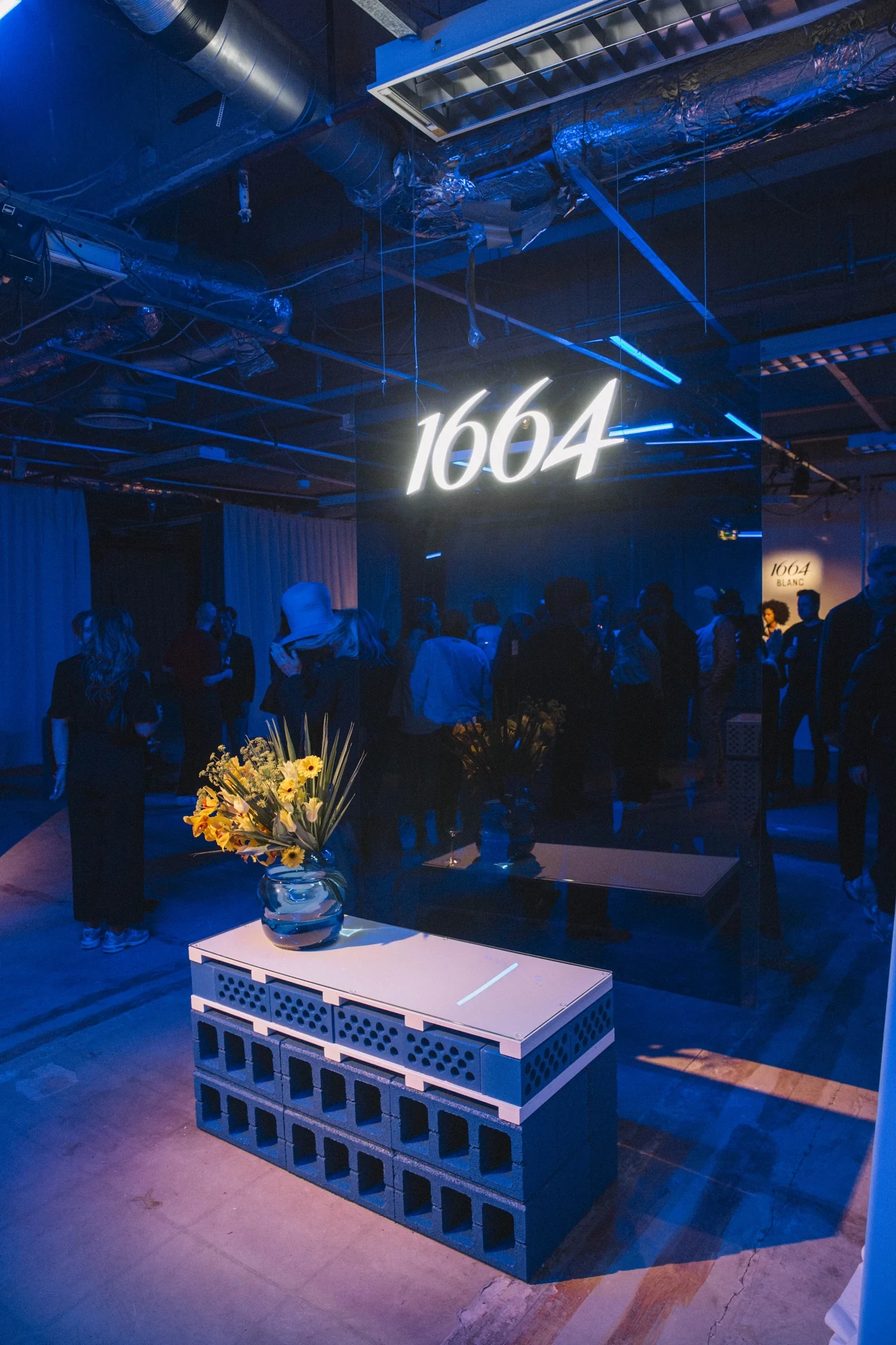 Event venue featuring a glowing neon „1664" sign suspended above a modular bar counter with sunflower arrangements, set in an industrial space bathed in deep blue light.