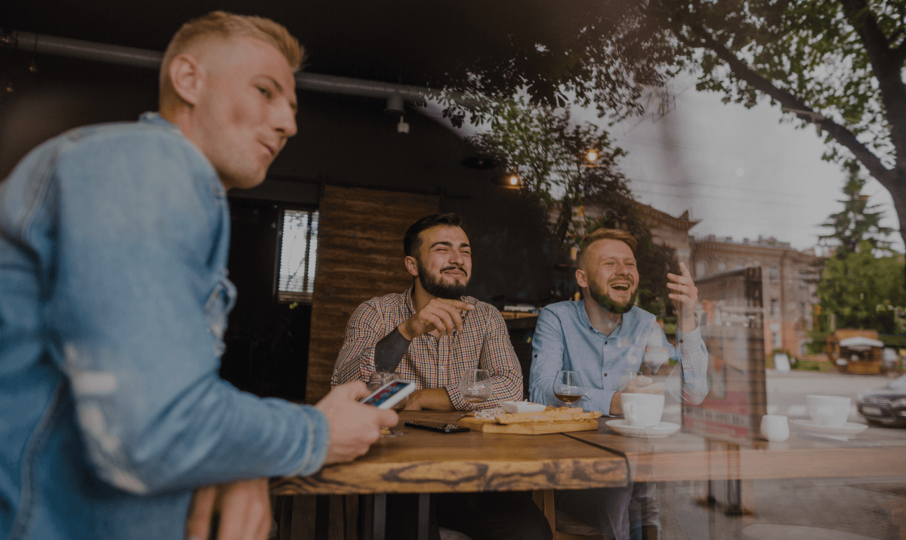 Una escena informal de tres amigos riendo y hablando mientras están sentados en una mesa de café. La foto fue tomada desde fuera, mirando a través de la ventana, capturando una atmósfera cálida y relajada. Una persona sostiene un teléfono, y hay comida y bebidas en la mesa.