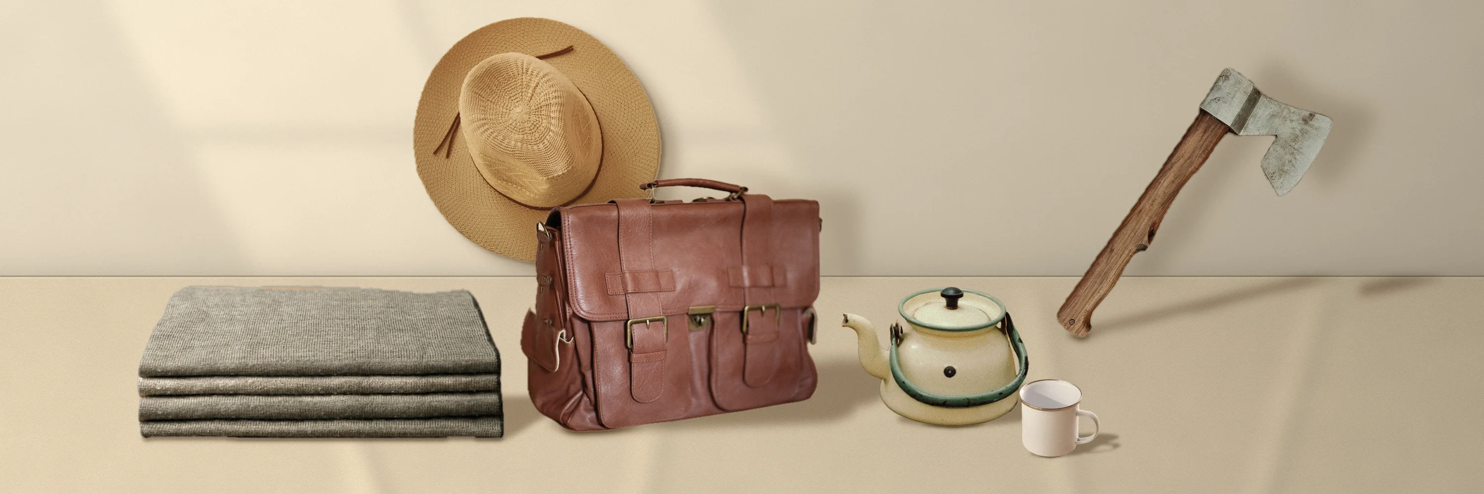 Leather satchel, enamel kettle, folded blanket, a straw hat and a hatchet on a sand-coloured table