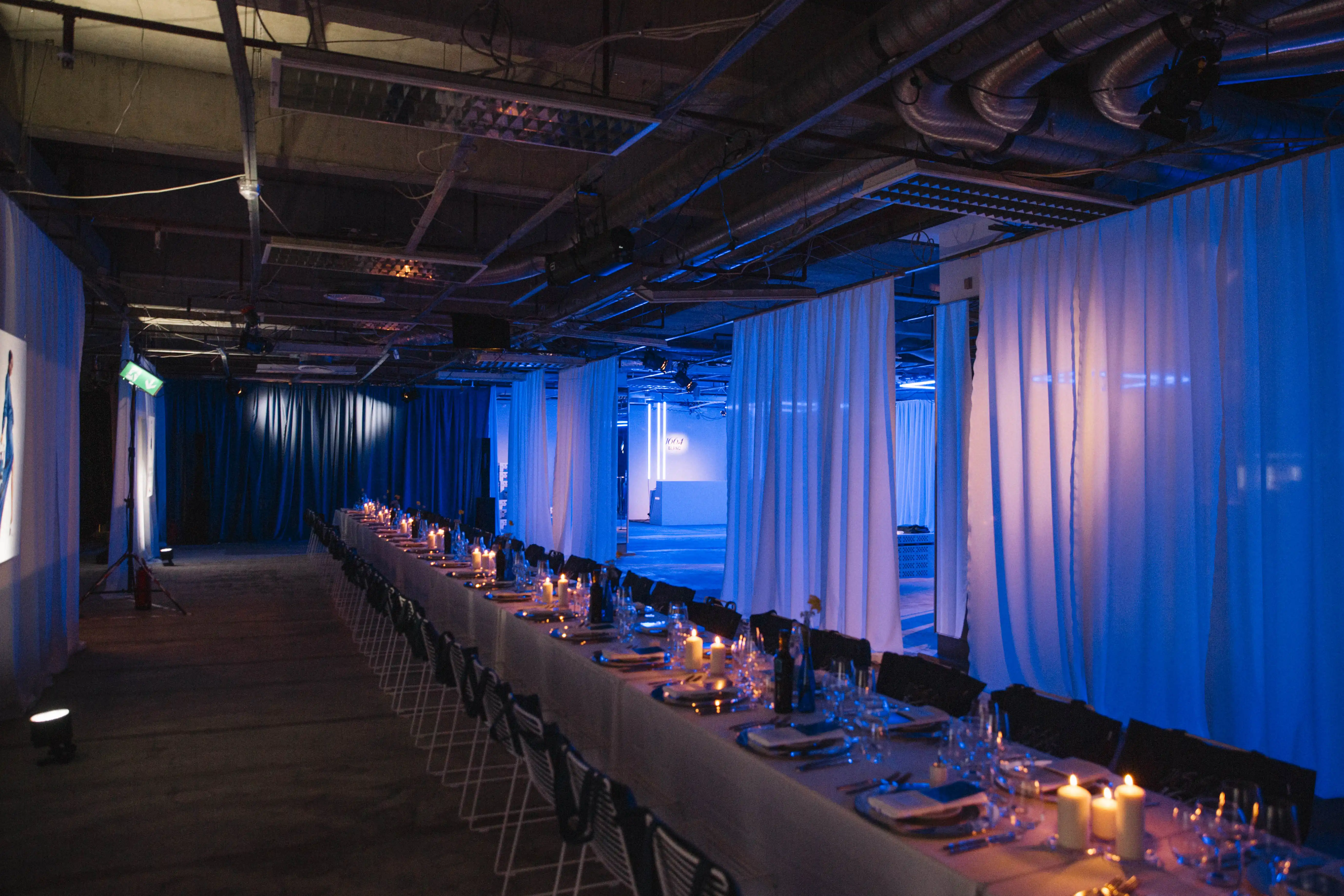 Long candle-lit dinner table between white curtains in the blue-lit industrial event space.