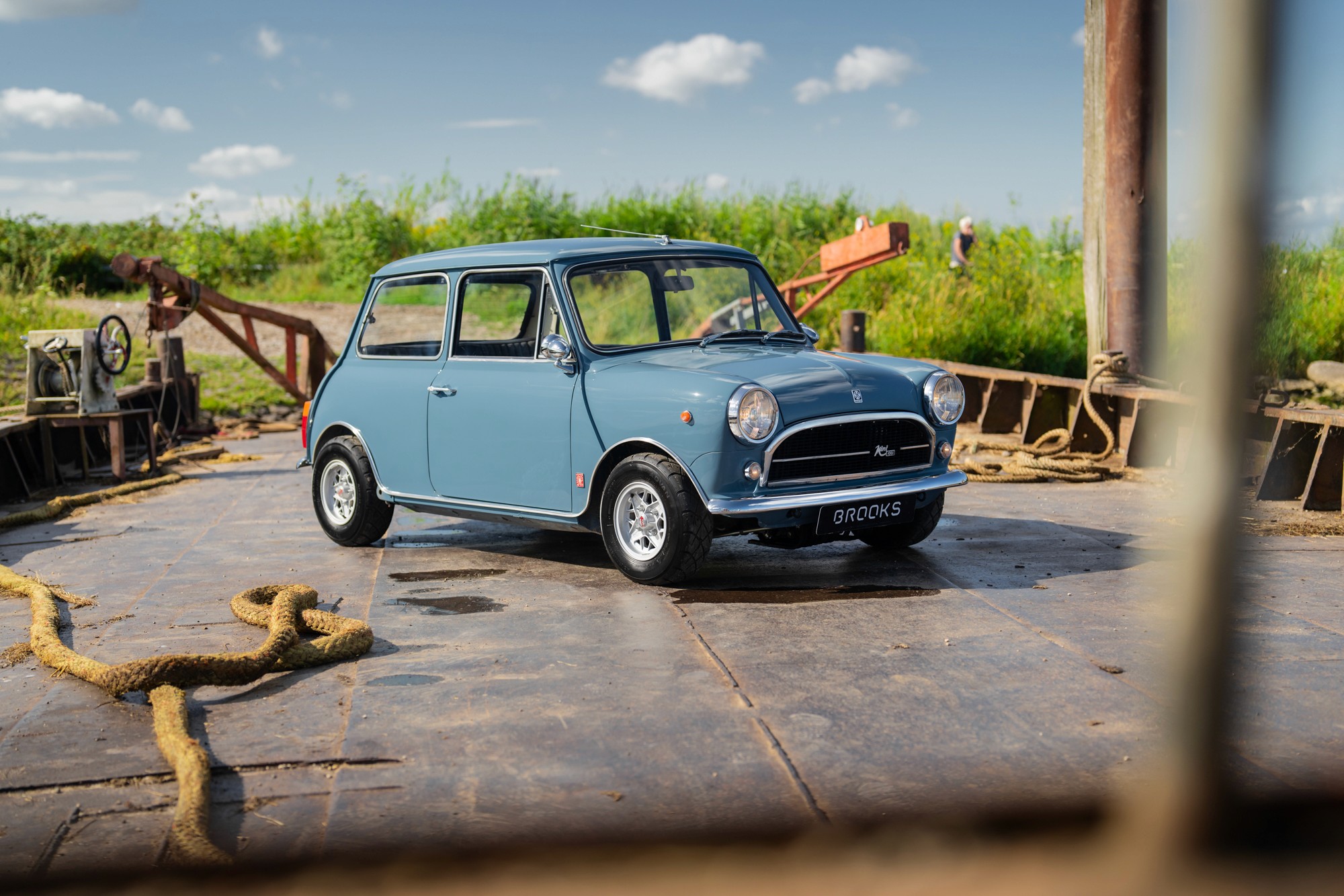 A vintage green mini classic automatic car drives along a road, surrounded by fields and a clear blue sky in the background.