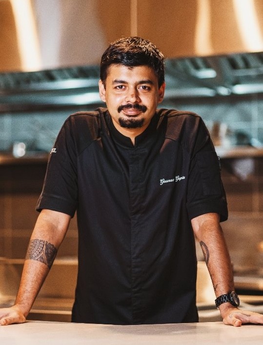 Chef Gaurav Gupta posing confidently in a kitchen environment, wearing a black chef's jacket with his name embroidered on it.