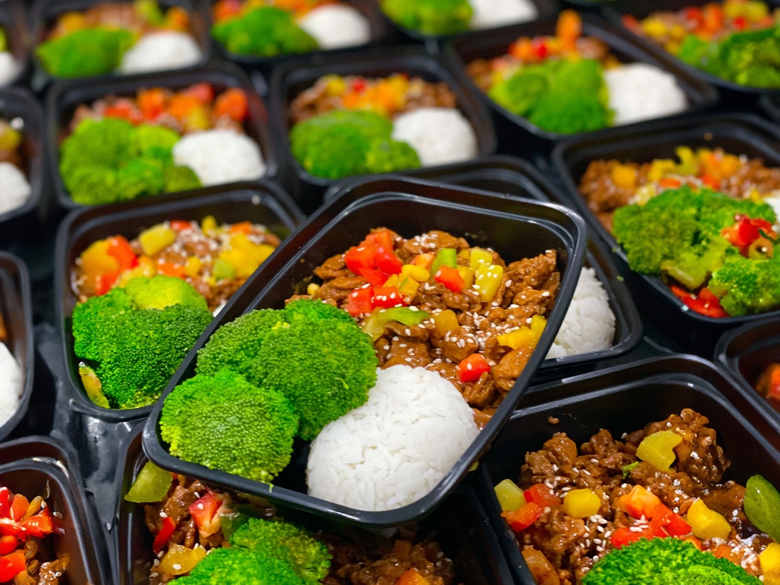 A display of prepared meals featuring colorful rice dishes with bright green broccoli arranged neatly in black containers, accompanied by a small white dipping bowl.