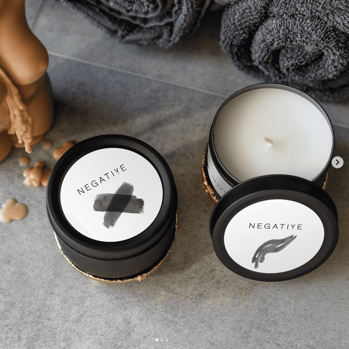 Two candles with black lids and decorative labels sit on a gray surface, surrounded by natural elements.