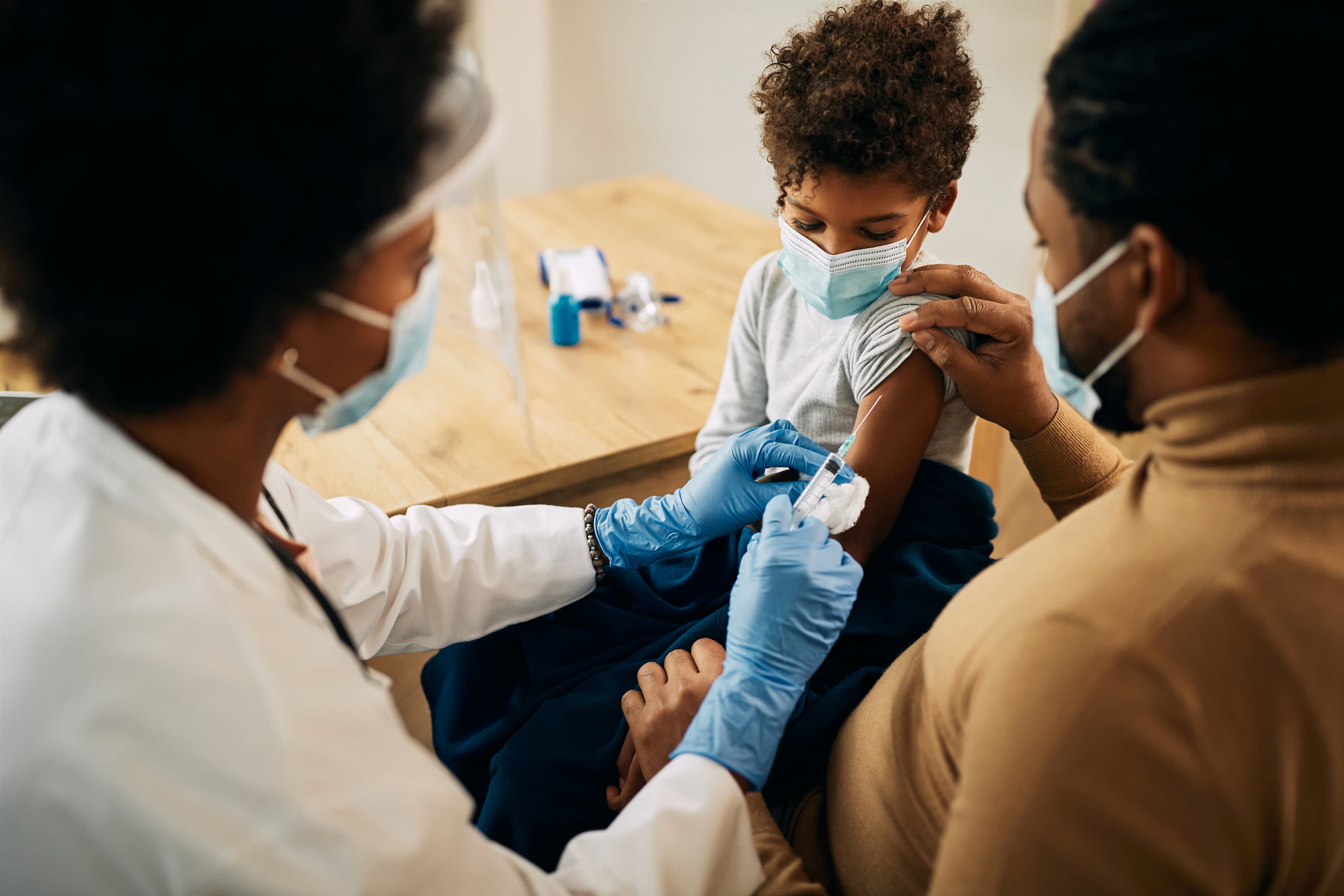 A healthcare worker administering a vaccine to a young boy wearing a mask while his parent gently holds his arm for support.