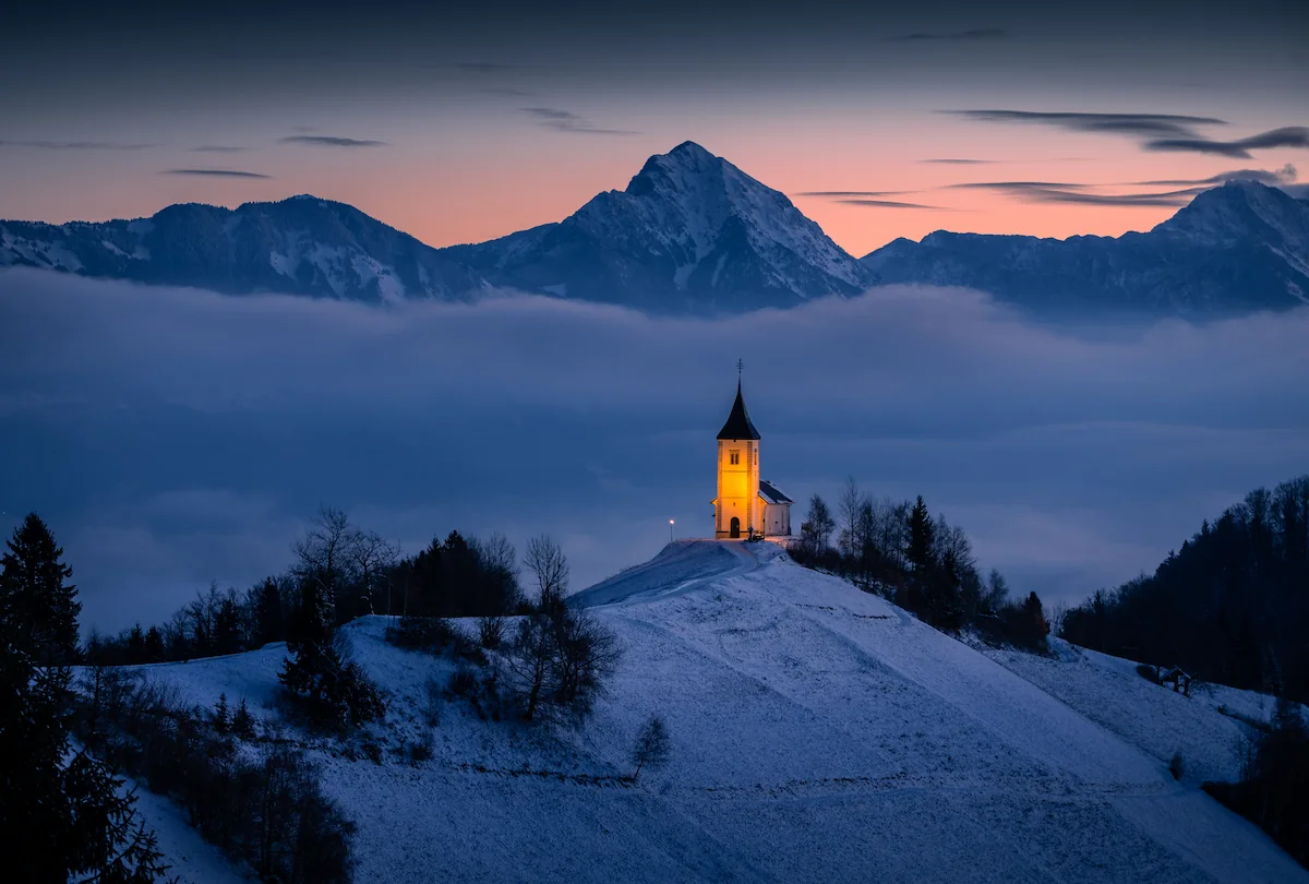 Church of St. Primus and Felician on Jamnik Hill, Slovenia, illuminated at twilight during winter, with the Kamnik-Savinja Alps and a sea of fog in the background.