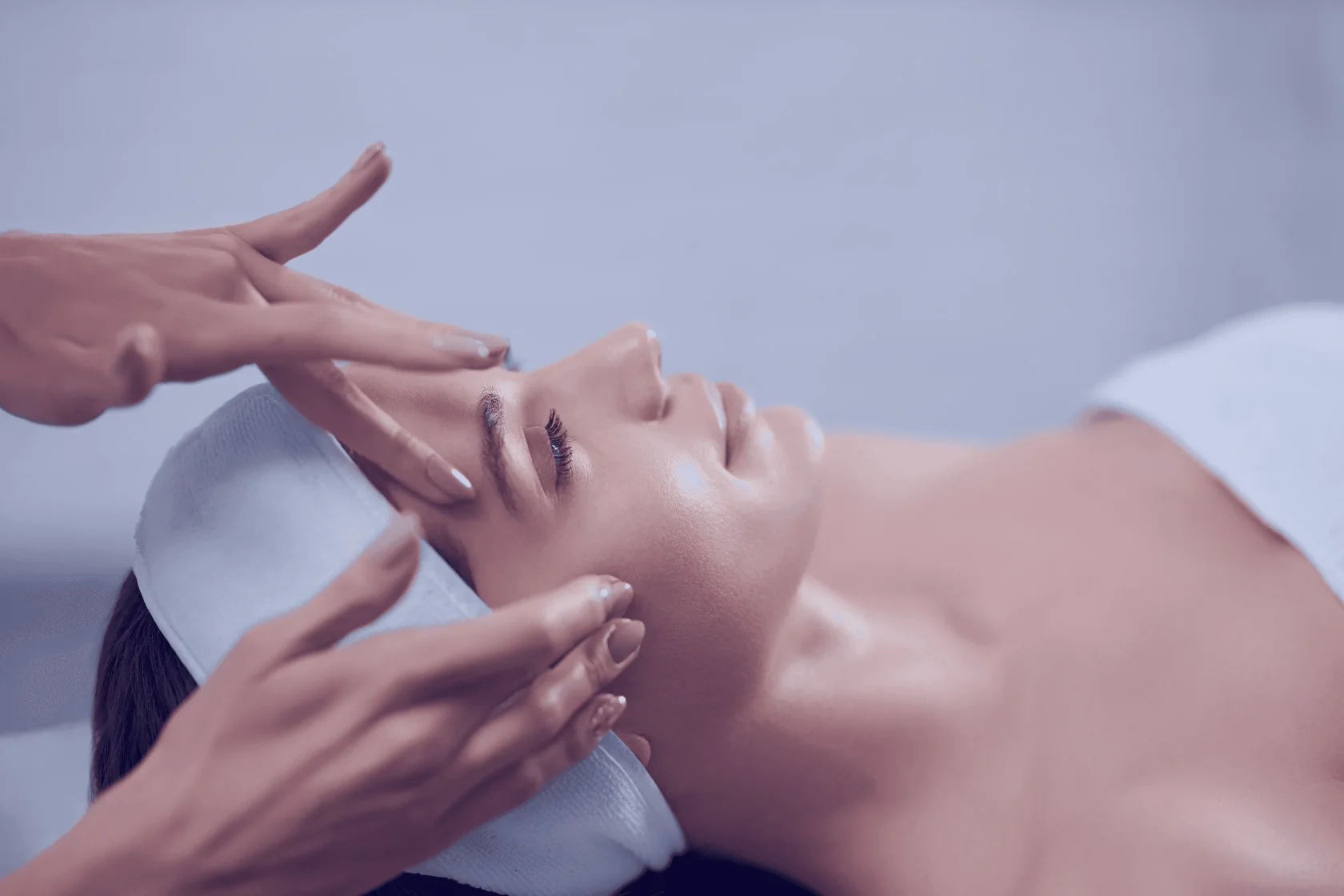 A person is lying on their back with closed eyes, receiving a relaxing facial in a peaceful spa setting, with hands gently massaging their forehead and a towel wrapped around their head.