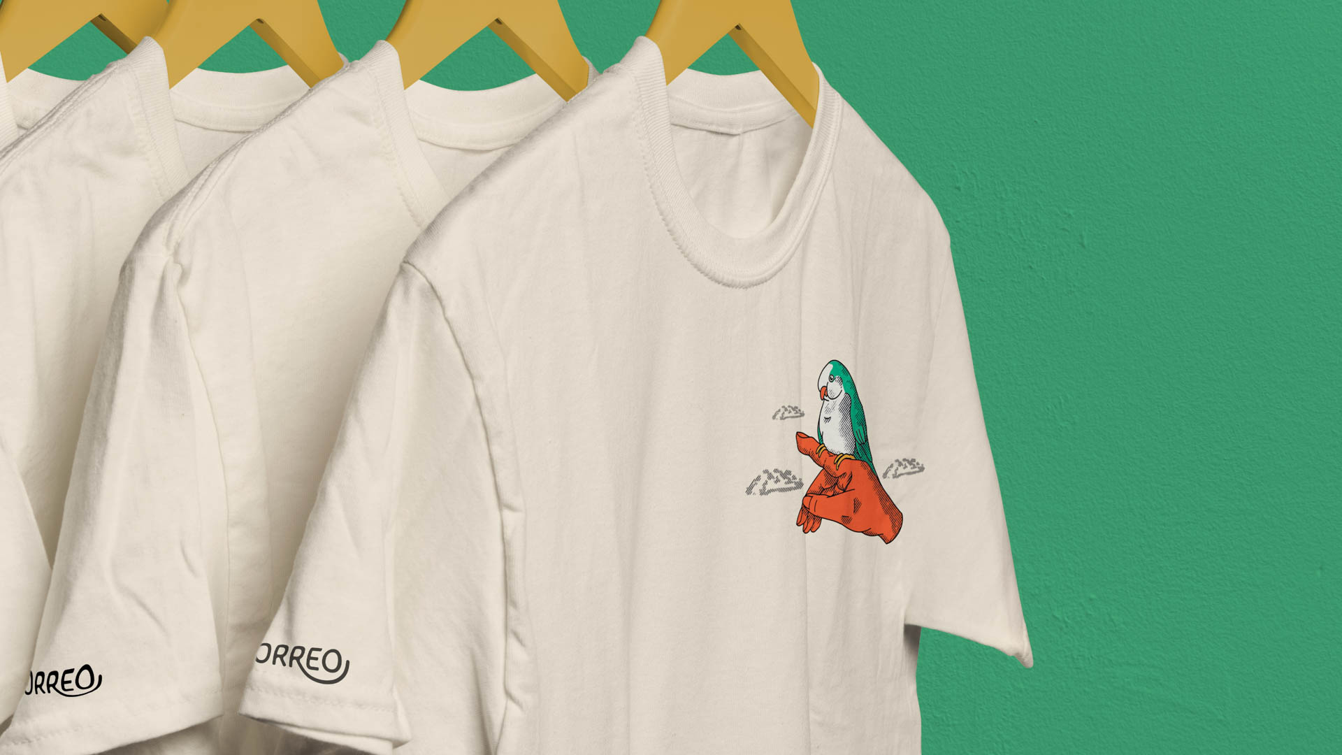 Three white t-shirts hang from wooden hangers against a teal wall. The shirt on the far left has a black stylized "ORREO" logo on the sleeve. The center shirt displays a cartoon bird in orange and green perched on a branch with small clouds. The shirt on the far right has no visible logo.