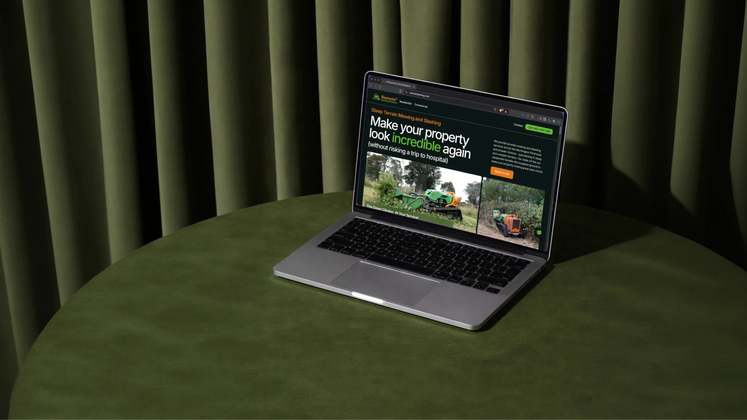 A silver laptop displaying a landscaping website on its screen is placed on a round, olive-green table with a background of dark green curtains.