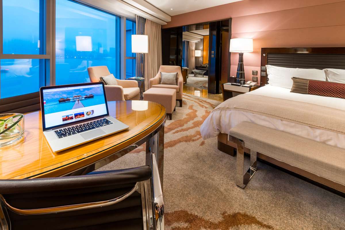 A modern cruise ship cabin with a large window overlooking the ocean, featuring a bed, two armchairs, and a laptop on a desk.
