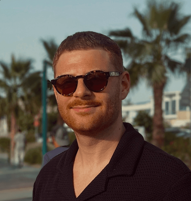 A person with curly hair and glasses smiles at the camera, seated in a cozy workspace with warm lighting.