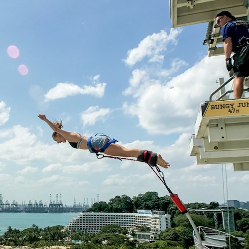 一个人在空中从平台上蹦极跳下，一名教练在旁边观看。下面可见海岸线风景和建筑物。