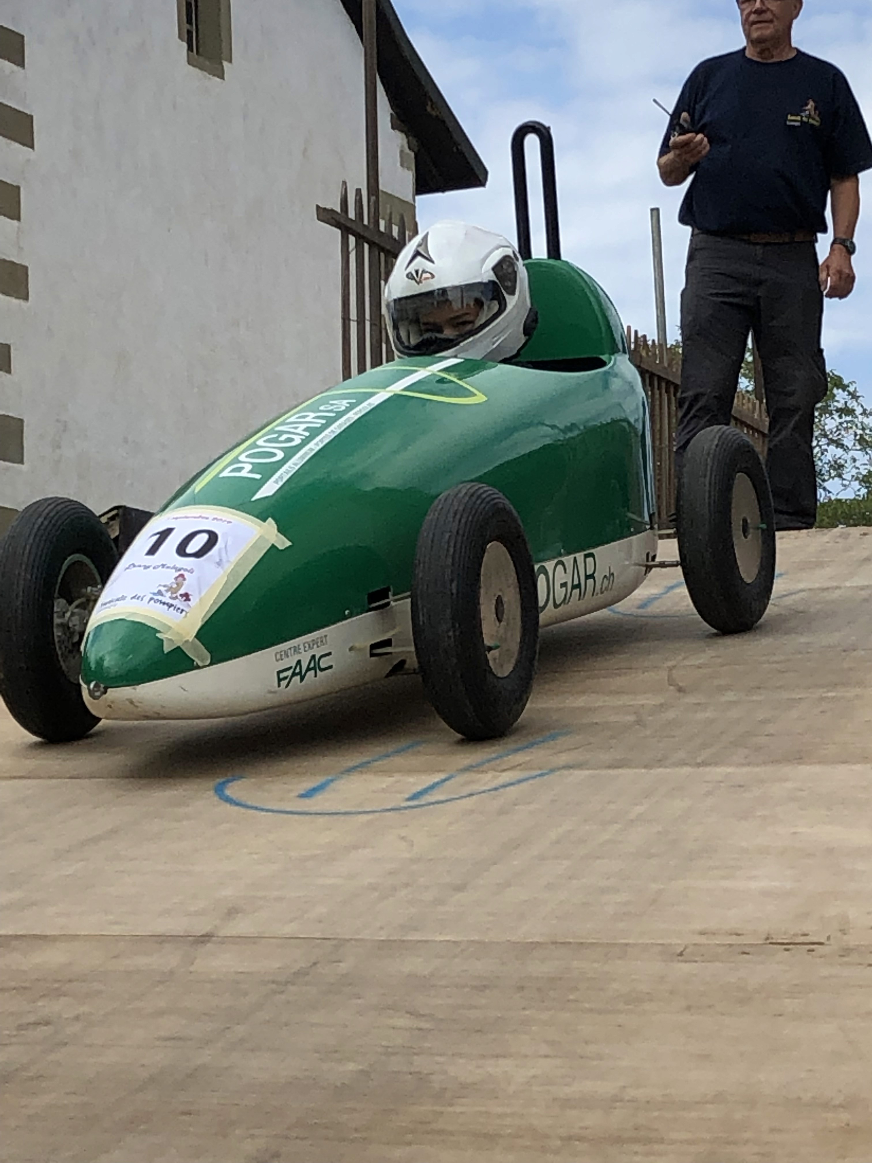 Petite voiture de course verte de type caisse à savon numéro 10, avec un enfant portant un casque blanc à l’intérieur, au sommet d’une rampe de départ, tandis qu’un adulte se tient derrière pour la guider."  Option un peu plus optimisée SEO / accessibilité :  "Caisse à savon verte