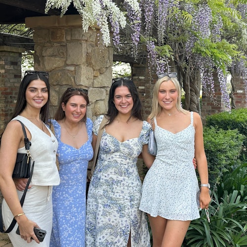 四名女性站在铺满紫色花朵的小径前。背景中可以看到Dave的巴士。