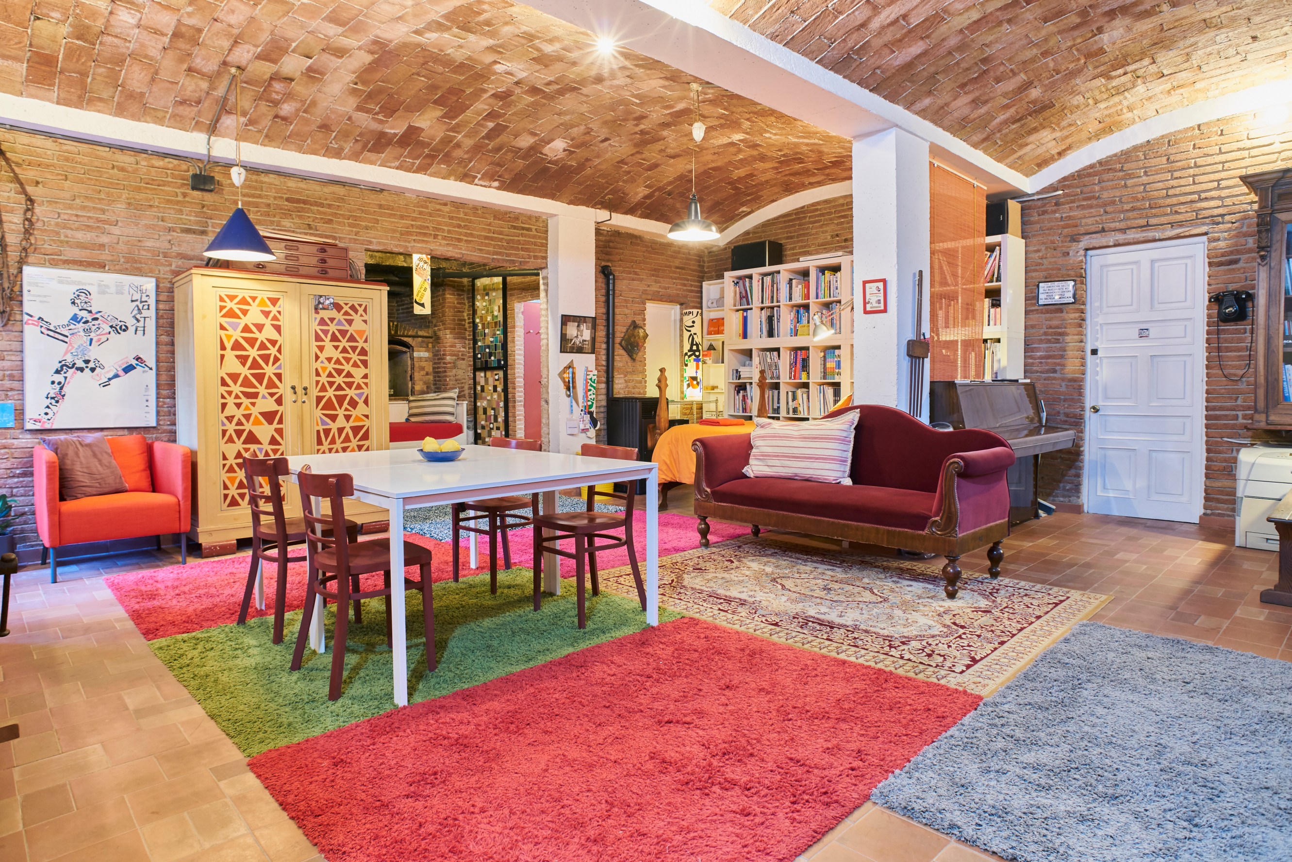 A cozy living space with arched brick ceilings, colorful rugs, a dining table with chairs, a red sofa, and a bookshelf filled with books.