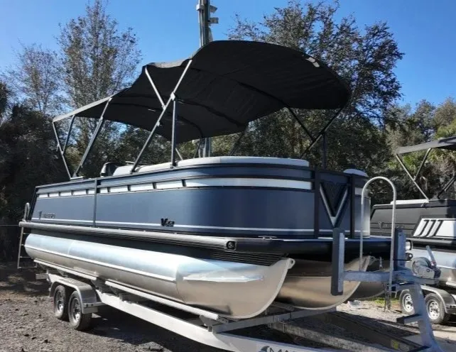 A sleek pontoon boat with a black canopy is parked on a trailer in a sunny outdoor setting in Bradenton, FL, surrounded by lush trees.
