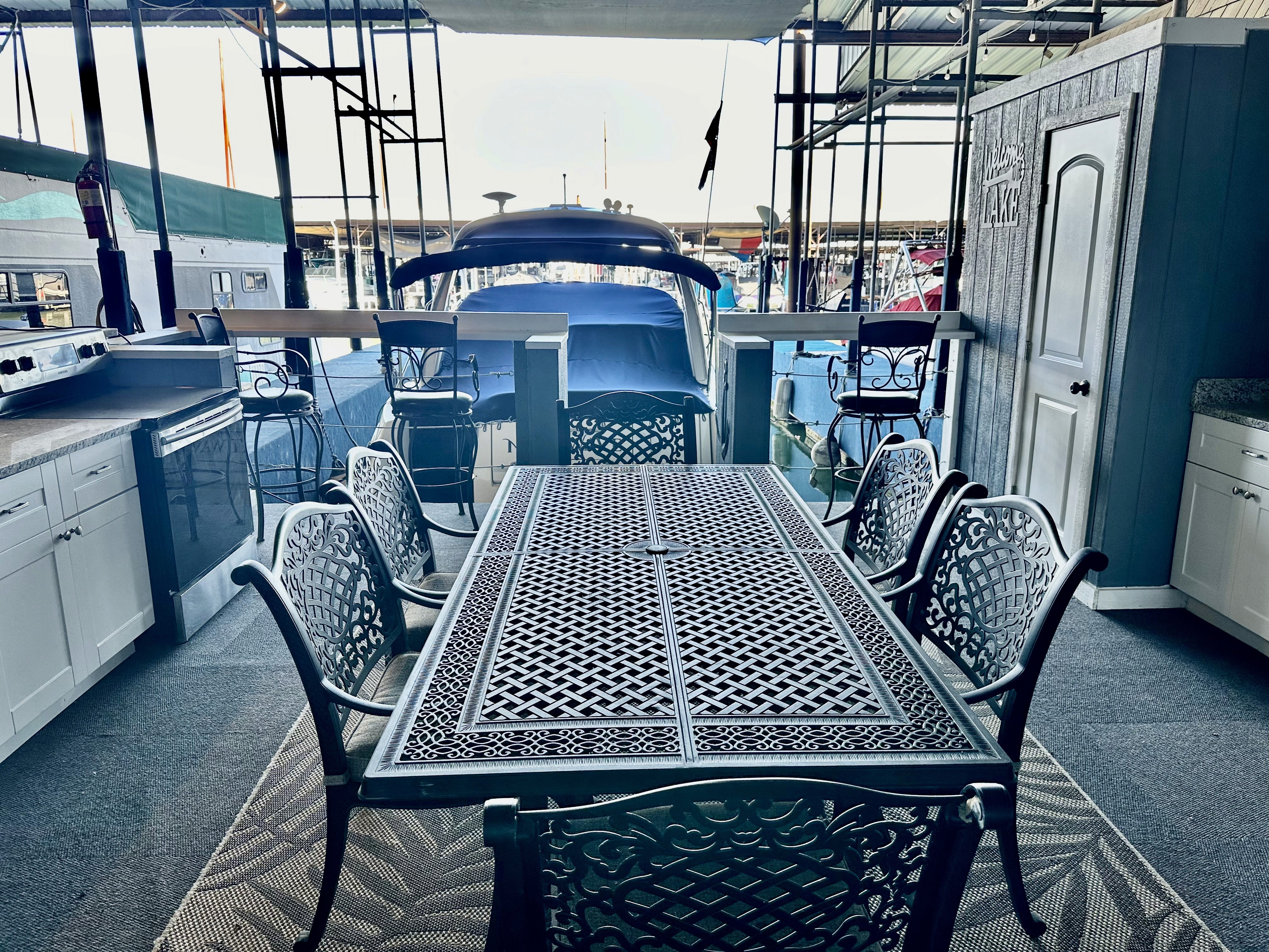 An elegantly set outdoor dining area features a long metal table with intricate designs, surrounded by matching chairs, all overlooking a docked boat in a marina; the space includes a kitchen setup with cabinets and refrigerators, conveying a luxurious marine lifestyle.
