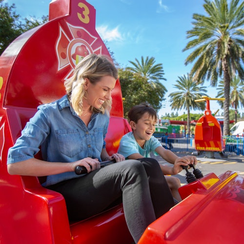 A woman and a child smile while seated on a bright red amusement ride with a firefighter theme. Palm trees are visible in the background.