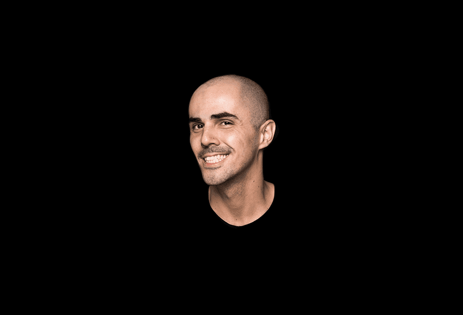 A man with a shaved head and a light complexion is shown against a solid black background. He is smiling, showing his teeth, and looking slightly to his right. He wears a dark-colored shirt.