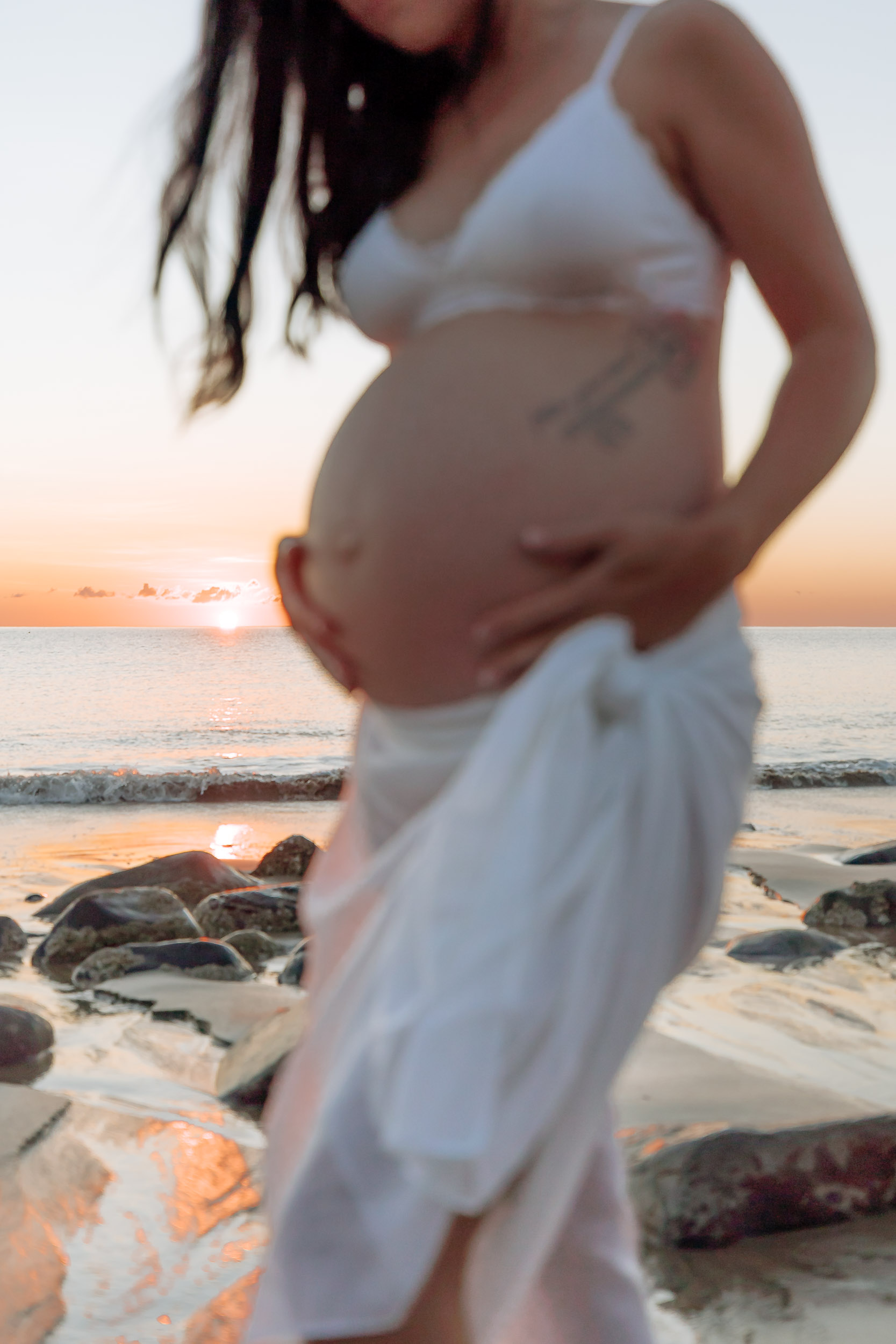 Beach maternity photoshoot at dawn featuring a pregnant woman silhouetted against the rising sun, natural light highlighting her baby bump and wind-swept hair.
