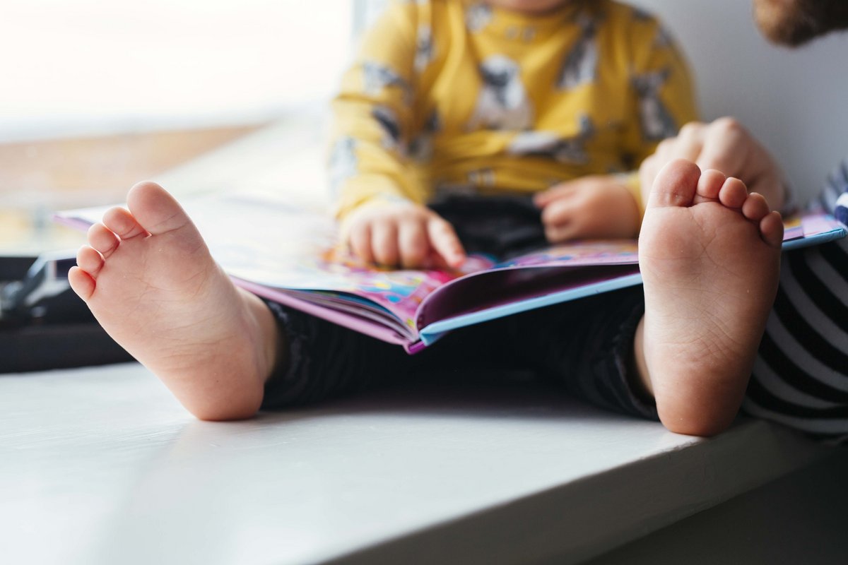 A child sits barefoot on a window sill, dressed in a yellow shirt with animal prints, reading a book on National Reading Day