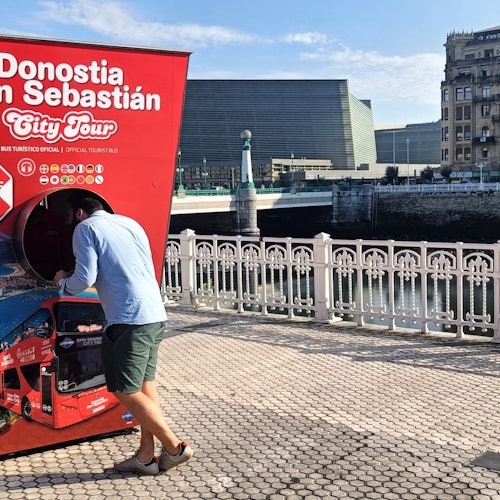 A red double-decker tour bus travels past a historic stone building with arched windows under a clear blue sky.