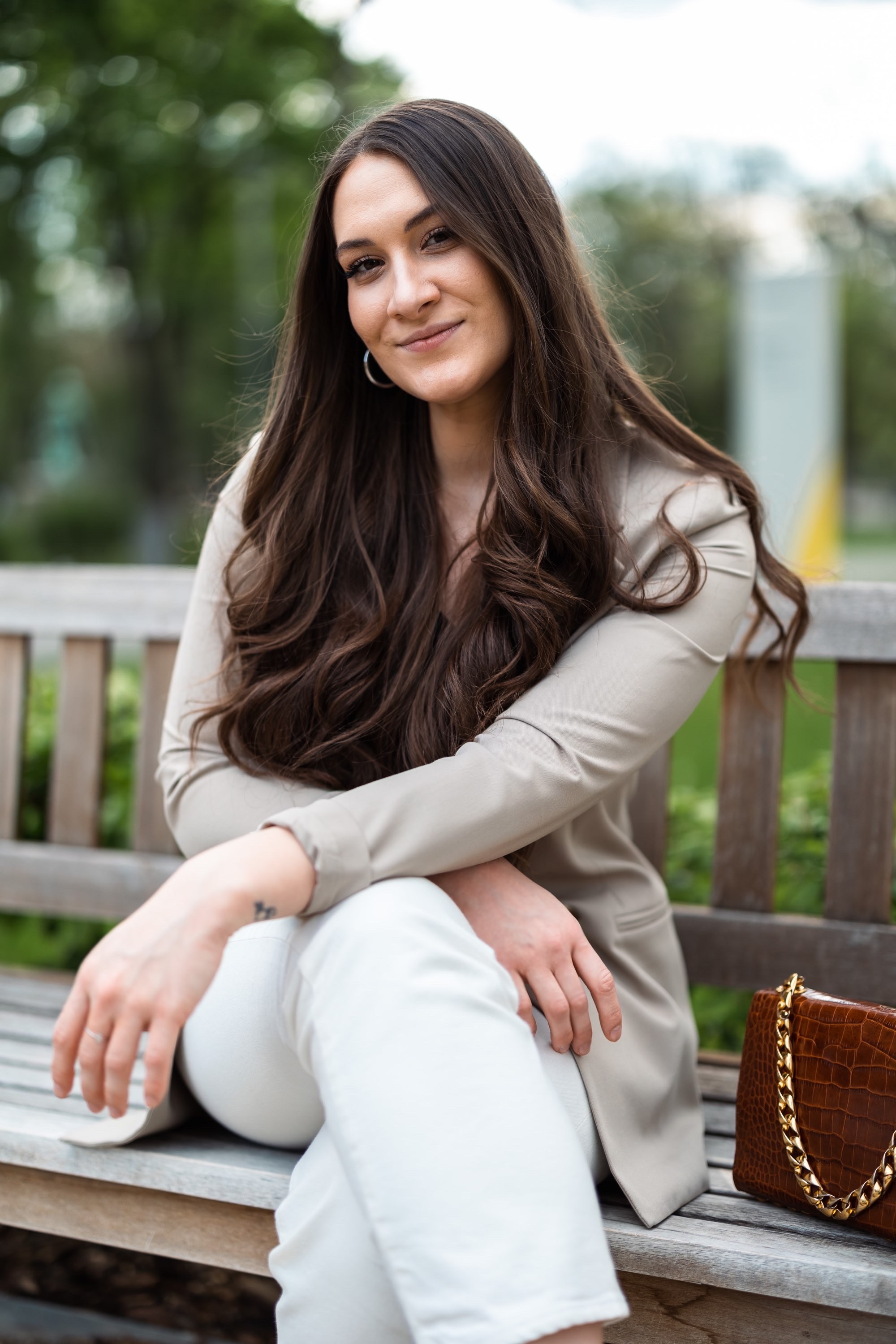 Fiatal nő oldalra tett lábbal, világos öltözékben, padon ülve, mellette barna táska, zöld parkban.