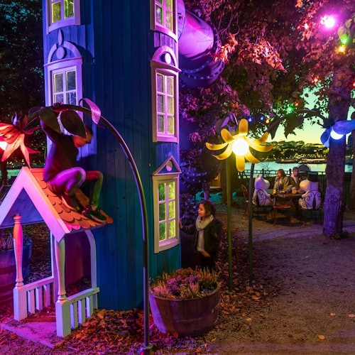 A whimsical outdoor scene with colorful lights, a blue playhouse, tree sculptures, and people at a table in the background.