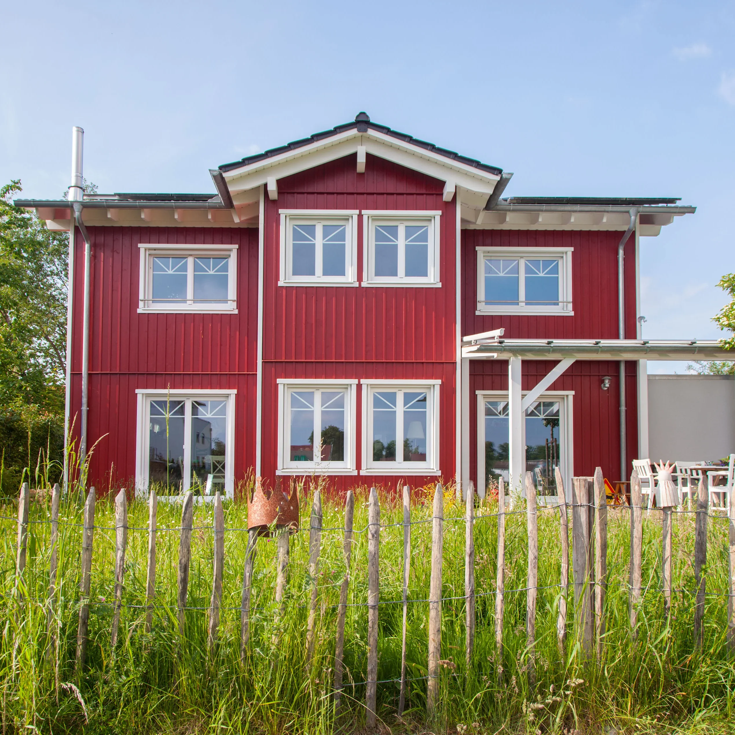 Ein rotes Holzhaus mit weißen Fenstern und Dachüberstand steht im Grünen hinter einem Holzzaun.